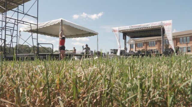 Personal técnico monta carpas y estructuras metálicas para el escenario del Alabaster CityFest en un campo de césped.