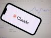 Anthropic lanza Claude Fable 5 con defensas de ciberseguridad Teléfono inteligente mostrando el logotipo de Anthropic Claude frente a una pantalla con gráficos financieros de bolsa.