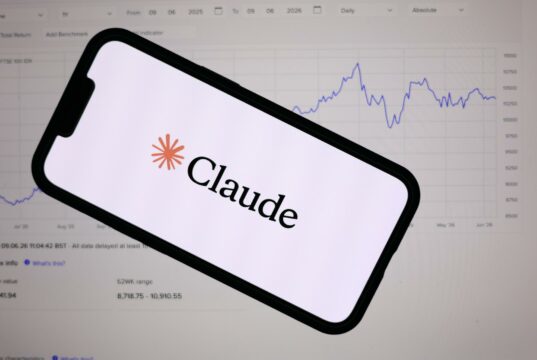 Anthropic lanza Claude Fable 5 con defensas de ciberseguridad Teléfono inteligente mostrando el logotipo de Anthropic Claude frente a una pantalla con gráficos financieros de bolsa.