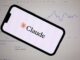 Anthropic lanza Claude Fable 5 con defensas de ciberseguridad Teléfono inteligente mostrando el logotipo de Anthropic Claude frente a una pantalla con gráficos financieros de bolsa.