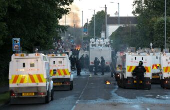 Disturbios en Belfast: Choques por el ataque de un refugiado Furgones blindados de la policía de Irlanda del Norte bloqueando una calle con humo y fuego de fondo durante los disturbios en Belfast.