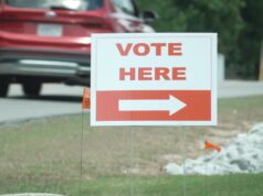 Elecciones en Alabama: Jefferson define su segunda vuelta Cartel blanco con letras rojas que dice 'Vote Here' junto a una flecha indicativa en una calle de Alabama.