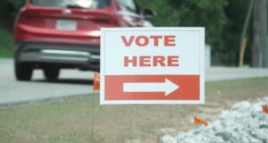 Elecciones en Alabama: Jefferson define su segunda vuelta Cartel blanco con letras rojas que dice 'Vote Here' junto a una flecha indicativa en una calle de Alabama.