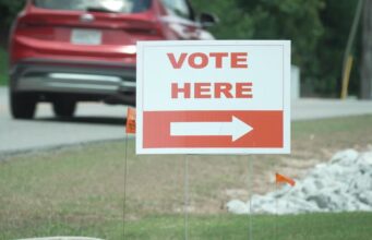 Elecciones en Alabama: Jefferson define su segunda vuelta Cartel blanco con letras rojas que dice 'Vote Here' junto a una flecha indicativa en una calle de Alabama.