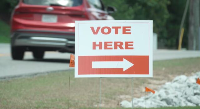 Elecciones en Alabama: Jefferson define su segunda vuelta Cartel blanco con letras rojas que dice 'Vote Here' junto a una flecha indicativa en una calle de Alabama.