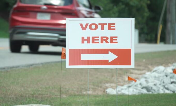 Elecciones en Alabama: Jefferson define su segunda vuelta Cartel blanco con letras rojas que dice 'Vote Here' junto a una flecha indicativa en una calle de Alabama.