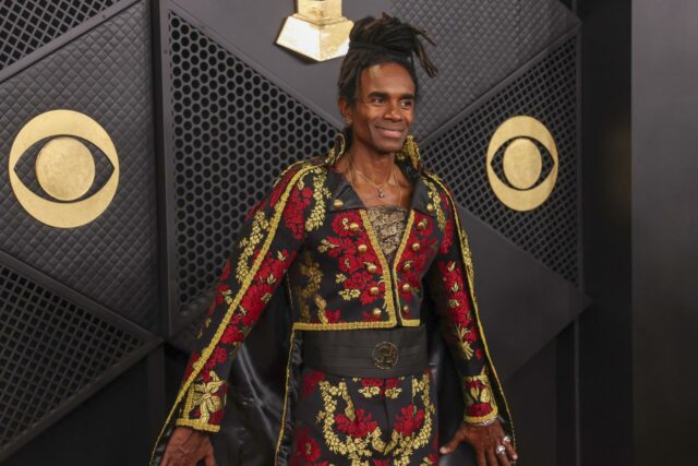 El cantante Fab Morvan posa sonriente en una alfombra roja con un traje negro y rojo bordado.