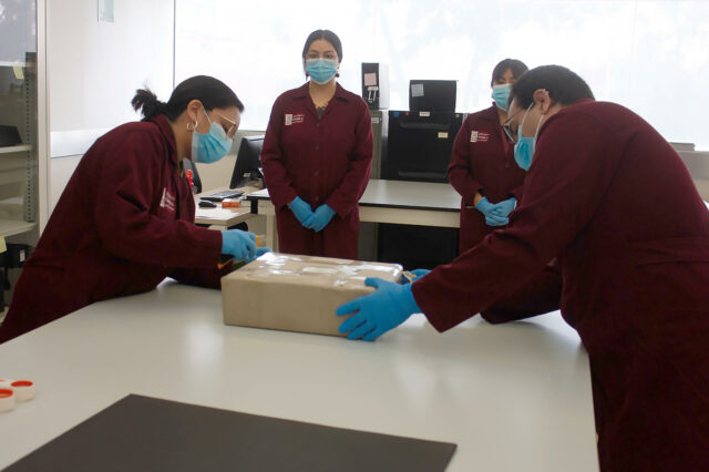 Personal calificado de México con batas guindas y guantes azules inspecciona una caja con un documento histórico.