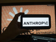 Modelos de IA en EE. UU. serán testados bajo nueva orden Mano sosteniendo un teléfono móvil con el logotipo de Anthropic frente a una pantalla de ordenador con la interfaz del software.