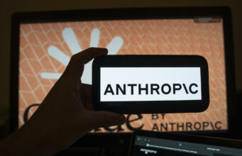 Modelos de IA en EE. UU. serán testados bajo nueva orden Mano sosteniendo un teléfono móvil con el logotipo de Anthropic frente a una pantalla de ordenador con la interfaz del software.