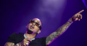 Gira de Ozuna 2026 arranca con espectacular concierto en Ibiza El cantante de reggaetón Ozuna cantando con micrófono y señalando hacia arriba con gafas de sol oscuras en concierto.