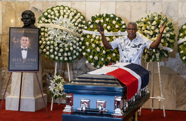 Capilla ardiente de Alex Bueno con su féretro cubierto por la bandera dominicana y arreglos florales en su honor.