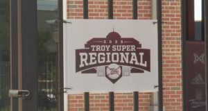 Súper Regionales: Troy aplasta a Little Rock en el Juego 1 Lona oficial con el logotipo del torneo Troy Super Regional 2026 colgada en una reja de ladrillos.