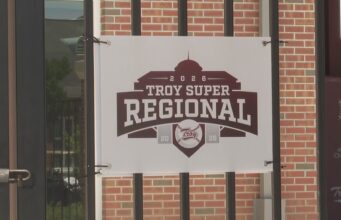 Súper Regionales: Troy aplasta a Little Rock en el Juego 1 Lona oficial con el logotipo del torneo Troy Super Regional 2026 colgada en una reja de ladrillos.