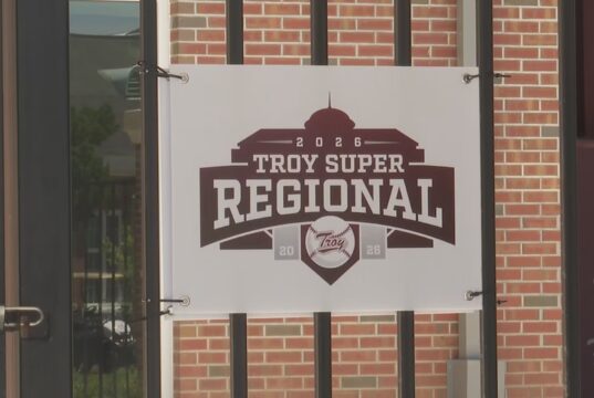 Súper Regionales: Troy aplasta a Little Rock en el Juego 1 Lona oficial con el logotipo del torneo Troy Super Regional 2026 colgada en una reja de ladrillos.
