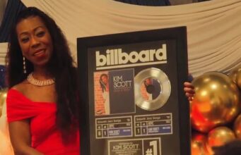 Kim Scott alcanza nuevo éxito número uno en Billboard La flautista Kim Scott sostiene un cuadro conmemorativo del número uno de Billboard por su canción Hide and Seek.