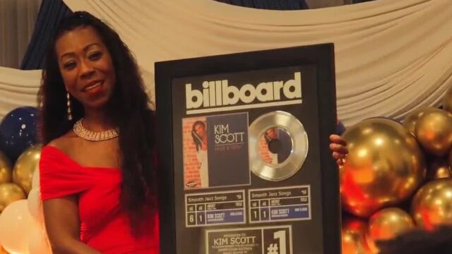 kim-scott-reconocimiento-billboard-sencillo-hide-and-seek La flautista Kim Scott sostiene un cuadro conmemorativo del número uno de Billboard por su canción Hide and Seek.