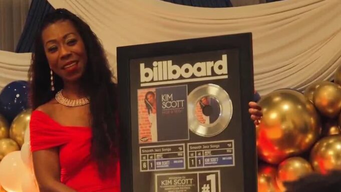 Kim Scott alcanza nuevo éxito número uno en Billboard La flautista Kim Scott sostiene un cuadro conmemorativo del número uno de Billboard por su canción Hide and Seek.