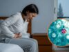 Alerta de salud por brote de ciclosporiasis y diarrea Mujer sentada en la cama expresando dolor abdominal intenso junto a micrografía del parásito Cyclospora cayetanensis.