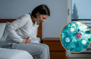Alerta de salud por brote de ciclosporiasis y diarrea Mujer sentada en la cama expresando dolor abdominal intenso junto a micrografía del parásito Cyclospora cayetanensis.