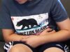 Alabama reporta 28 casos de Cyclospora en brote nacional Niño sentado en un sofá con expresión de dolor abdominal, indicando síntomas de ciclosporiosis.