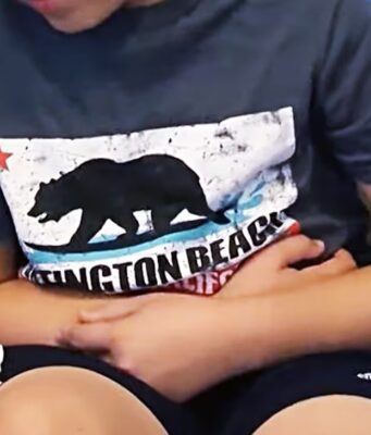 Alabama reporta 28 casos de Cyclospora en brote nacional Niño sentado en un sofá con expresión de dolor abdominal, indicando síntomas de ciclosporiosis.