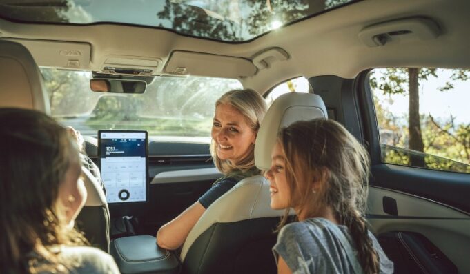 Vehículos eléctricos tendrán pruebas de manejo gratis Una madre sonríe a sus hijas desde el asiento delantero de un moderno vehículo eléctrico con techo panorámico de cristal.