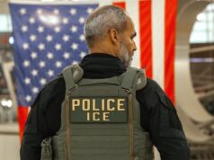 Guantes eléctricos para el ICE desatan fuerte rechazo Agente del ICE de espalda con chaleco táctico frente a la bandera de Estados Unidos.
