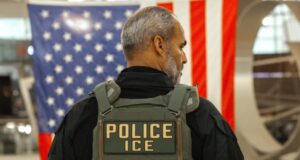 Guantes eléctricos para el ICE desatan fuerte rechazo Agente del ICE de espalda con chaleco táctico frente a la bandera de Estados Unidos.