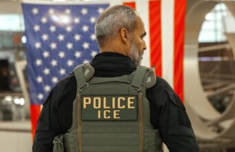 Guantes eléctricos para el ICE desatan fuerte rechazo Agente del ICE de espalda con chaleco táctico frente a la bandera de Estados Unidos.