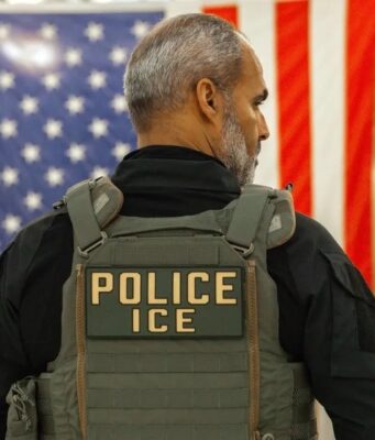 Guantes eléctricos para el ICE desatan fuerte rechazo Agente del ICE de espalda con chaleco táctico frente a la bandera de Estados Unidos.