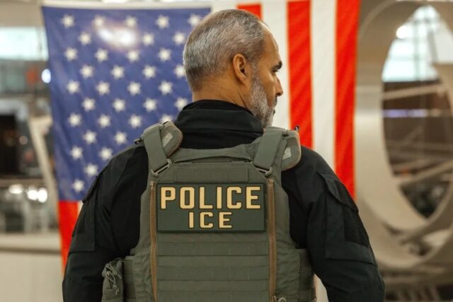 Agente del ICE de espalda con chaleco táctico frente a la bandera de Estados Unidos.