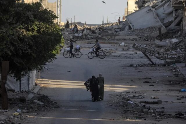 Civiles y ciclistas recorren una calle rodeada de escombros y edificios destruidos tras ataques en Gaza.