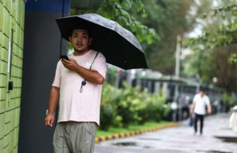 China declara alertas rojas por lluvias e inundaciones Hombre en China sosteniendo un paraguas negro bajo la lluvia mientras mira su teléfono móvil.