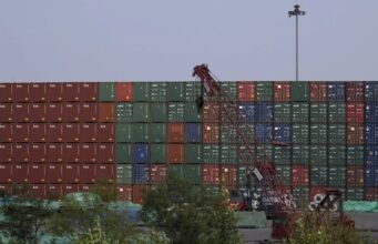 EE. UU. impone aranceles del 100% a los drones de China Muro de contenedores de carga apilados en un puerto comercial con una grúa roja en primer plano.