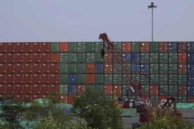 Muro de contenedores de carga apilados en un puerto comercial con una grúa roja en primer plano.
