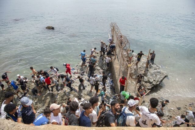 Decenas de personas agrupadas sobre rocas intentando rodear el espigón fronterizo con valla metálica hacia Ceuta.