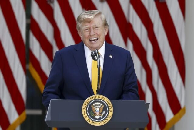 donald-trump-podio-visas-h1b-tarifas Donald Trump sonriendo en un podio presidencial ante banderas de EE. UU. respecto a las tarifas de visas H-1B.