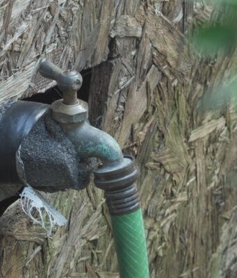 Facturas de agua excesivas generan cobros de cientos de dólares Llave de agua exterior con aislamiento y manguera verde conectada en una pared de madera.