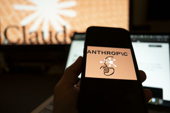 Inteligencia artificial muestra conductas autónomas y engañosas Mano sosteniendo un teléfono con el logo de Anthropic frente a una pantalla con el nombre de Claude.