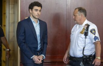 Luigi Mangione se declara culpable por crimen de Brian Thompson Luigi Mangione en traje formal y esposado junto a un oficial dentro de un tribunal de Nueva York.