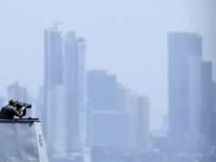 EE. UU. lanza fuerza multinacional de drones de ataque Oficial de la Marina de EE. UU. en la cubierta de un buque observando el horizonte frente a un perfil urbano.