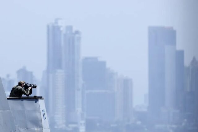 Oficial de la Marina de EE. UU. en la cubierta de un buque observando el horizonte frente a un perfil urbano.