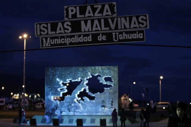 plaza-islas-malvinas-ushuaia-tierra-del-fuego Letrero nocturno de la Plaza Islas Malvinas en la Municipalidad de Ushuaia con un monumento iluminado al fondo.
