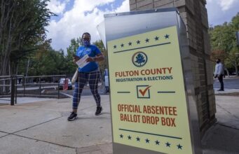 Justicia de EE. UU. respalda restricciones al voto por correo Buzón de entrega de voto por correo de Fulton County en Estados Unidos con una votante caminando al fondo.