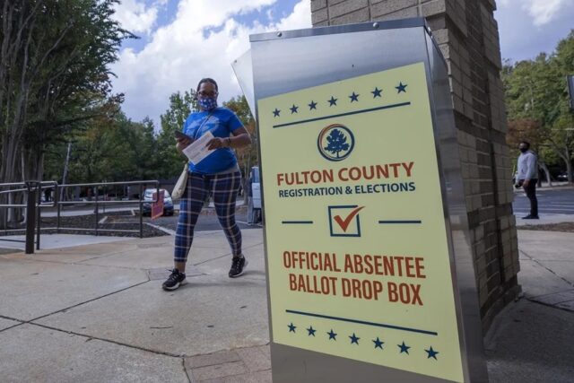 Buzón de entrega de voto por correo de Fulton County en Estados Unidos con una votante caminando al fondo.