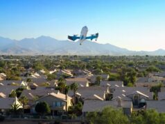 Amazon expandirá entrega con drones Prime Air a 500 ciudades Dron de entrega de Amazon Prime Air volando sobre un vecindario residencial con montañas al fondo.