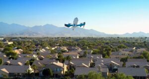 Amazon expandirá entrega con drones Prime Air a 500 ciudades Dron de entrega de Amazon Prime Air volando sobre un vecindario residencial con montañas al fondo.