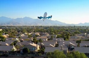 Amazon expandirá entrega con drones Prime Air a 500 ciudades Dron de entrega de Amazon Prime Air volando sobre un vecindario residencial con montañas al fondo.