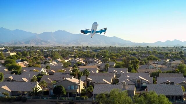 dron-prime-air-amazon-vuelo-residencial Dron de entrega de Amazon Prime Air volando sobre un vecindario residencial con montañas al fondo.